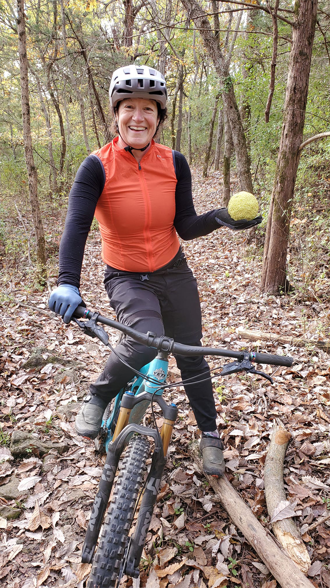 Berne Broudy on her mountain bike on a wooded trail, holding an osage orange